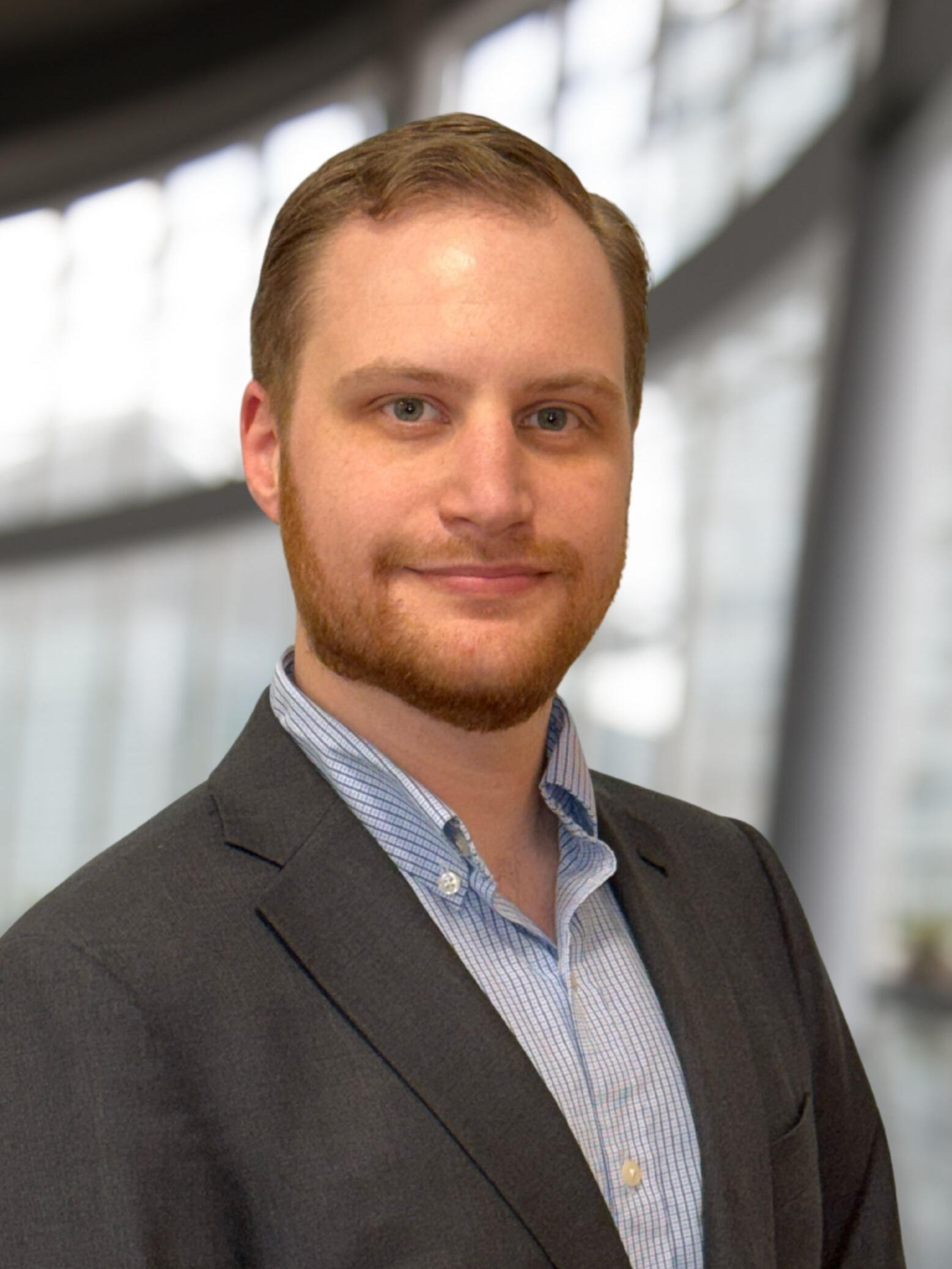 Professional portrait of Michael Bartolomeo in a dark suit jacket and a light blue collared shirt, looking directly at the camera with a slight smile. The background is a blurred, grayscale modern office or building interior.